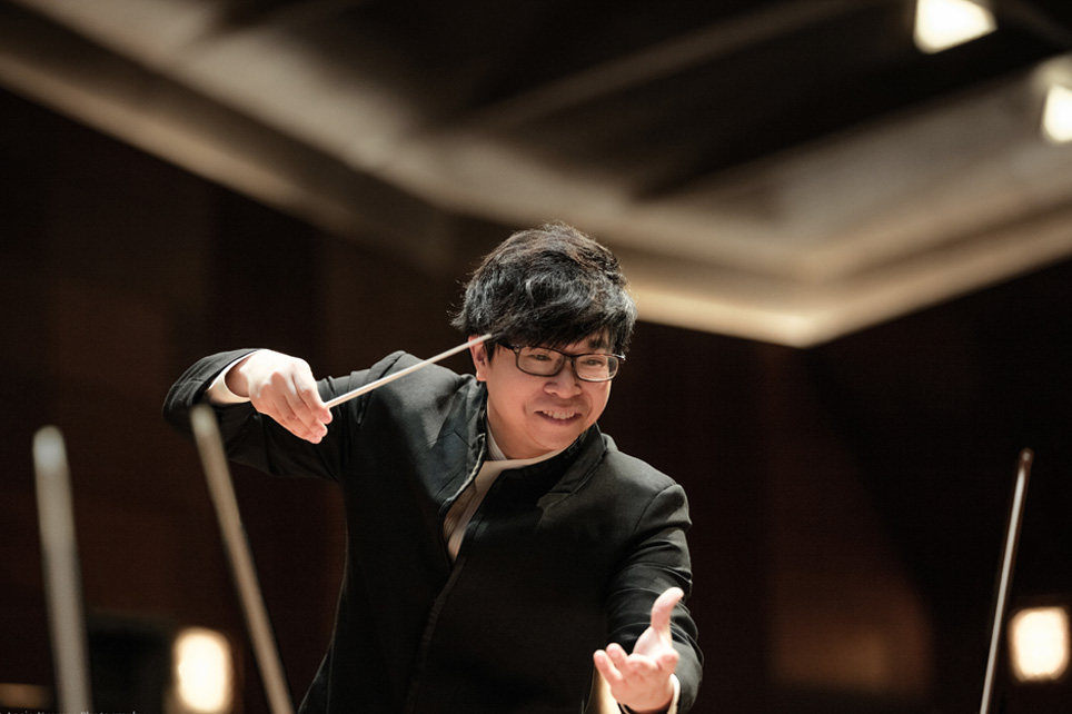 Kahchun Wong, a man wearing smart black attire, conducting at the Queen Elizabeth Hall, smiling in a performance. Kahchun Wong, a man wearing smart black attire, conducting at the Queen Elizabeth Hall, smiling in a performance.
